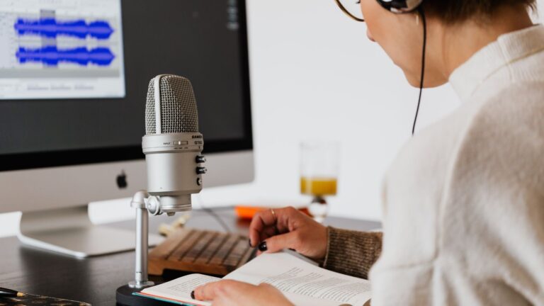 Eine Person mit Kopfhörer spricht in ein Mikrofon vor einem Computerbildschirm mit Audiobearbeitungssoftware und hält ein Notizbuch bereit.